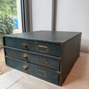 Peut inclure: Un meuble de rangement à trois tiroirs de couleur bleu sarcelle foncé avec une finition usée. Chaque tiroir est doté d'un petit bouton rond en métal foncé et d'un porte-étiquette rectangulaire en laiton. Le meuble est rectangulaire.