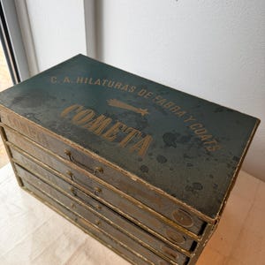 Può includere: Un mobiletto rettangolare verde scuro a quattro cassetti, con la scritta C. A. HILATURAS DE FABRA Y COATS e COMETA e un grafico a stella cadente stampato in oro sulla superficie superiore. Ogni cassetto è dotato di una piccola maniglia in metallo.