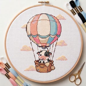 Peut inclure: Broderie au point de croix d'une vache dans une montgolfière. Le ballon est rayé de rose, de bleu et de blanc. La vache est noire et blanche, dans un panier en osier. La broderie est dans un cercle en bois, avec du fil à broder et des ciseaux à proximité.