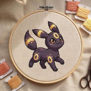Peut inclure: Broderie au point de croix d'un personnage de dessin animé Umbreon dans un cercle en bois. L'Umbreon est principalement violet foncé avec des accents jaunes. Le fond est un tissu beige clair. Des bobines de fil orange et jaune sont visibles.