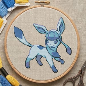 Peut inclure: Un projet de broderie au point de croix représentant une créature pixelisée bleue et blanche avec de grandes oreilles et une longue queue, présenté dans un tambour en bois circulaire sur un tissu beige clair, avec du fil jaune et bleu.