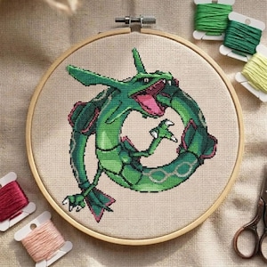 May include: A cross-stitch embroidery of a green dragon with pink accents, set in a wooden hoop. The dragon is in a circular pose, with its mouth open. Spools of green, pink, and peach thread are arranged around the hoop.
