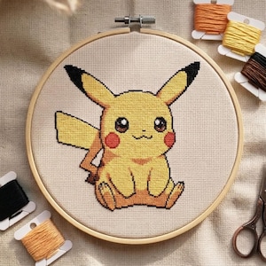 Peut inclure: Broderie au point de croix de Pikachu, un personnage Pokémon jaune, dans un cercle en bois. Le motif représente Pikachu avec des oreilles à pointes noires, des joues rouges et une expression souriante. Des bobines de fil et des ciseaux sont visibles.