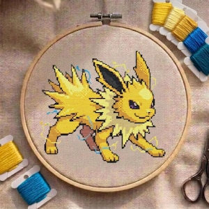 Peut inclure: Un projet de broderie au point de croix dans un tambour en bois circulaire représentant une créature jaune à quatre pattes avec une fourrure hérissée, des oreilles pointues et des éclairs bleus. Le motif est brodé sur un tissu beige clair.