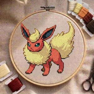 Peut inclure: Un projet de broderie au point de croix dans un tambour en bois circulaire représentant une créature stylisée avec une fourrure rouge, une grande crinière jaune et une queue touffue jaune. Le motif est brodé sur un tissu beige clair.