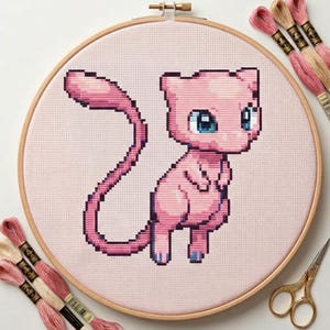 Peut inclure: Broderie au point de croix d'un personnage de chat rose de dessin animé aux yeux bleus, dans un cercle en bois. Le chat a une longue queue. Du fil à broder rose et crème et de petits ciseaux dorés sont également visibles.