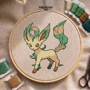 Peut inclure: Un projet de broderie au point de croix dans un tambour en bois circulaire représentant une créature stylisée avec une fourrure crème, des oreilles en forme de feuilles vertes et une queue touffue sur un tissu beige clair avec du fil.