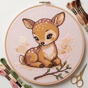 Peut inclure: Un cercle de broderie au point de croix représentant un faon avec des taches marron et blanches, de grands yeux bruns et un fond rose. La broderie est dans un cercle en bois, avec du fil à broder et des ciseaux à proximité. Le faon est couché avec une brindille de feuilles.