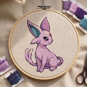 Peut inclure: Broderie au point de croix d'une créature de dessin animé violette dans un cercle en bois. La créature a de grandes oreilles avec des accents turquoise et une longue queue. Une gemme rouge est sur son front. Des bobines de fil et des ciseaux sont également visibles.