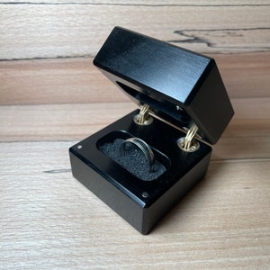 Può includere: Una scatola per anelli in legno nero con un coperchio incernierato, aperta per rivelare un anello d'argento che riposa su un cuscino di velluto nero.