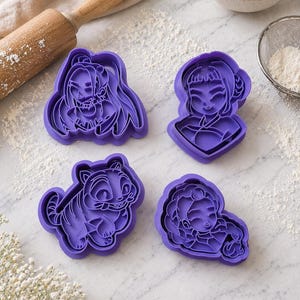 Könnte beinhalten: Vier violette Ausstecher in verschiedenen Formen: eine Frau mit langen Haaren, eine Frau mit Dutt, ein Tiger und eine Frau mit Zöpfen. Die Ausstecher liegen auf einer weißen Oberfläche mit Mehl und einer Teigrolle im Hintergrund. Back- und Küchenutensilien.