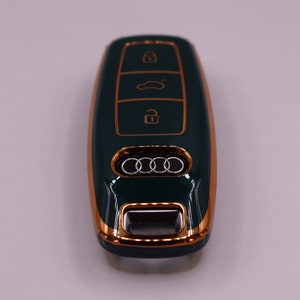 Puede incluir: Una llave de coche verde oscuro con un borde dorado y el logotipo de Audi. La llave tiene tres botones: bloquear, desbloquear y maletero.