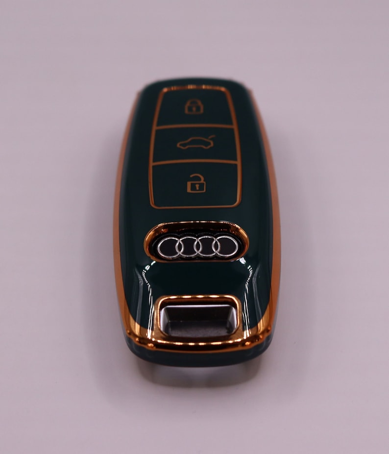 Puede incluir: Una llave de coche verde oscuro con un borde dorado y el logotipo de Audi. La llave tiene tres botones: bloquear, desbloquear y maletero.