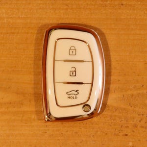 May include: A white and rose gold car key fob cover featuring a glossy finish. The front panel displays three icons: a locked padlock, an unlocked padlock, and a car trunk symbol with the text HOLD printed underneath in a dark font.