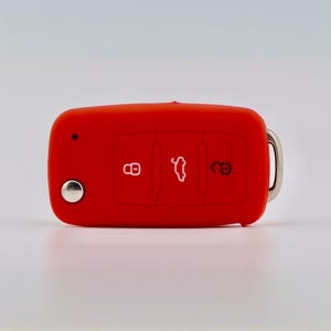 May include: A bright red silicone car key fob cover featuring three embossed icons: a locked padlock, a car trunk symbol, and an unlocked padlock. The cover includes a circular silver metal button on the left side and a silver metal ring on the right.