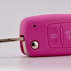 May include: A bright pink silicone car key fob cover featuring three embossed buttons with white and red icons for locking, unlocking, and trunk release. The metallic key blade is extended from the side of the rectangular, rounded-edge casing.