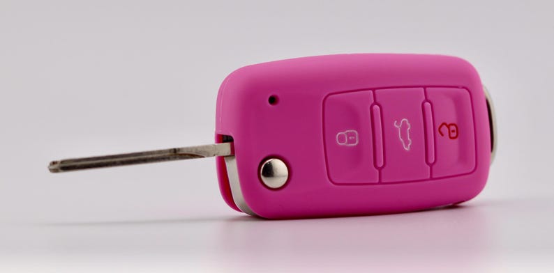 May include: A bright pink silicone car key fob cover featuring three embossed buttons with white and red icons for locking, unlocking, and trunk release. The metallic key blade is extended from the side of the rectangular, rounded-edge casing.