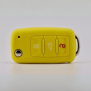 May include: A bright yellow silicone car key fob cover featuring three rectangular buttons. The left button displays a white lock icon, the center button shows a white car trunk icon, and the right button features a red unlocked padlock icon.