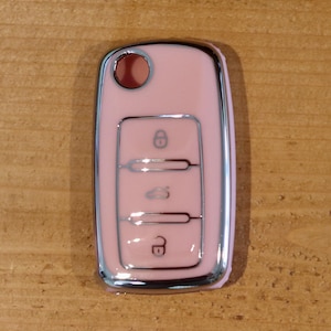 May include: A light pink car key fob cover with a glossy finish and a metallic silver border. The cover features three distinct button icons including a locked padlock, a car trunk symbol, and an unlocked padlock, with a circular hole at the top.