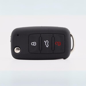 May include: A black rectangular car key fob featuring three rubberized buttons. The left button displays a white padlock icon, the center button shows a white car trunk icon, and the right button features a red open padlock icon. A silver metal button is on the left.