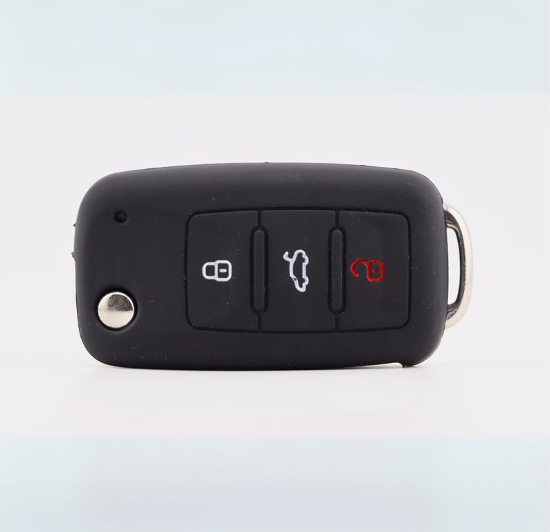 May include: A black rectangular car key fob featuring three rubberized buttons. The left button displays a white padlock icon, the center button shows a white car trunk icon, and the right button features a red open padlock icon. A silver metal button is on the left.