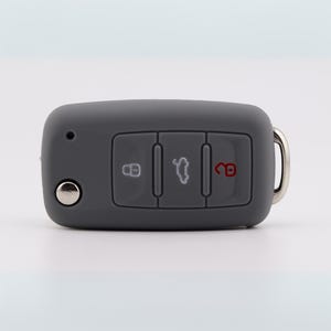 May include: A matte gray silicone car key fob cover featuring three rectangular buttons. The left button displays a white lock icon, the center button shows a white car trunk icon, and the right button features a red unlocked padlock icon.