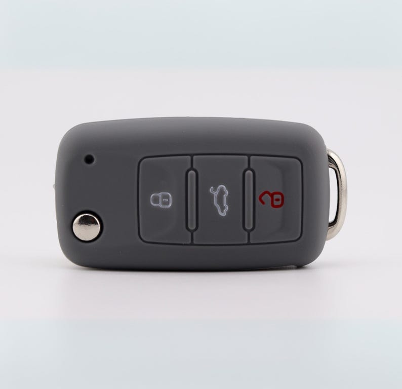 May include: A matte gray silicone car key fob cover featuring three rectangular buttons. The left button displays a white lock icon, the center button shows a white car trunk icon, and the right button features a red unlocked padlock icon.