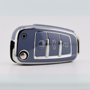 May include: A glossy slate blue car key fob cover featuring a metallic silver trim and three distinct buttons. The buttons display white icons representing a locked padlock, a car trunk, and an unlocked padlock, with a circular silver button.