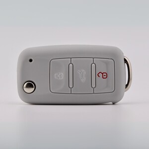 May include: A light gray silicone car key fob cover featuring three rectangular button cutouts. The left button displays a lock icon, the center button shows a trunk release icon, and the right button features an unlocked padlock icon in red.