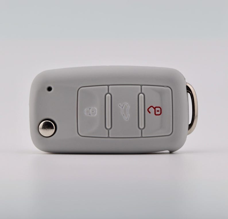 May include: A light gray silicone car key fob cover featuring three rectangular button cutouts. The left button displays a lock icon, the center button shows a trunk release icon, and the right button features an unlocked padlock icon in red.