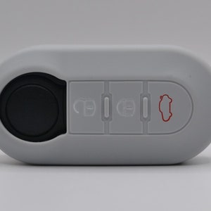 May include: White car key fob with four buttons. The buttons have icons for unlock, lock, trunk, and a car silhouette. The key fob has a black circular button on the left side.