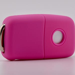 May include: A bright pink, rectangular-shaped electronic key fob cover with rounded edges. It features a circular cutout on the front surface and a silver-toned metal ring attached to one side for securing keys or other small items.
