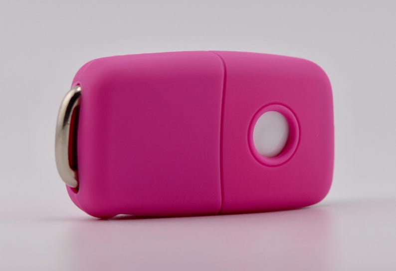 May include: A bright pink, rectangular-shaped electronic key fob cover with rounded edges. It features a circular cutout on the front surface and a silver-toned metal ring attached to one side for securing keys or other small items.