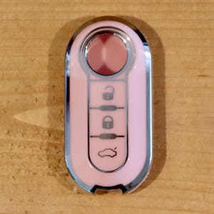 May include: A light pink car key fob with a metallic silver border. The device features a circular button at the top and three vertical rectangular buttons below, each marked with black icons representing an unlock, lock, and trunk release function.
