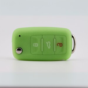 May include: A lime green silicone car key fob cover featuring three embossed buttons with white and red icons for locking, unlocking, and trunk release. The rectangular cover has a small circular cutout for a key blade and a metal ring attachment.