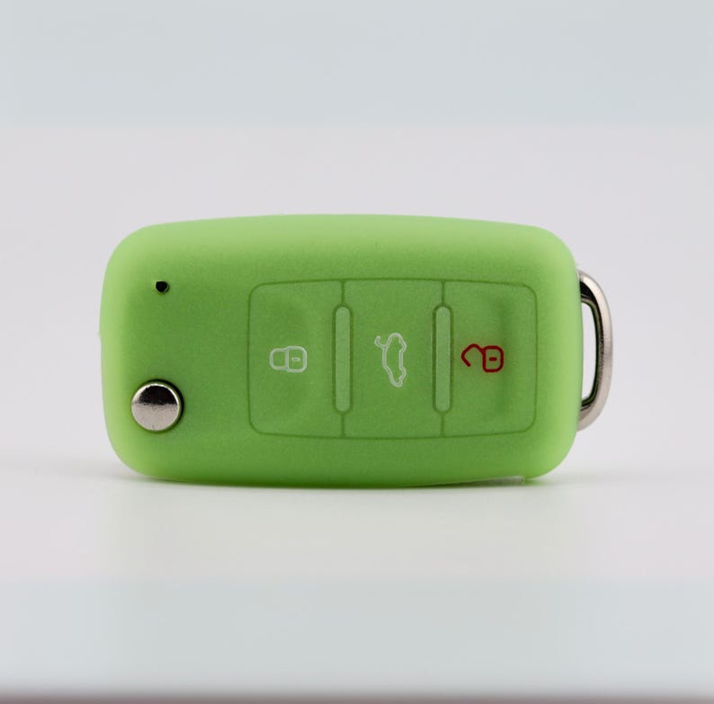 May include: A lime green silicone car key fob cover featuring three embossed buttons with white and red icons for locking, unlocking, and trunk release. The rectangular cover has a small circular cutout for a key blade and a metal ring attachment.