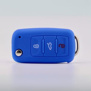 May include: A bright blue silicone car key fob cover featuring three recessed buttons with white icons for locking, trunk release, and unlocking. The cover has a small circular cutout on the left side and a silver metal ring attached to the right side.