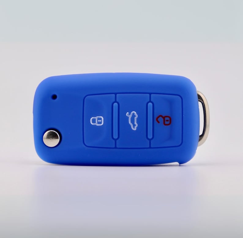 May include: A bright blue silicone car key fob cover featuring three recessed buttons with white icons for locking, trunk release, and unlocking. The cover has a small circular cutout on the left side and a silver metal ring attached to the right side.