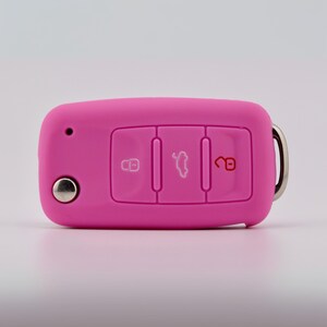 May include: A bright pink silicone car key fob cover featuring three embossed buttons with white and red icons for locking, unlocking, and trunk release. The rectangular cover includes a small circular cutout for a key blade and a metal ring attachment.