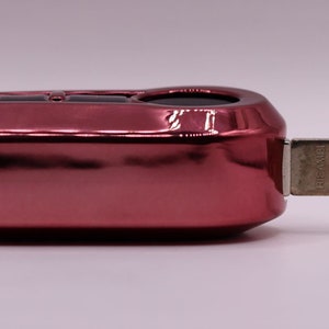 Puede incluir: Una llave de coche roja con una hoja de llave de metal plateado. La llave tiene un bot&oacute;n negro en la parte superior.