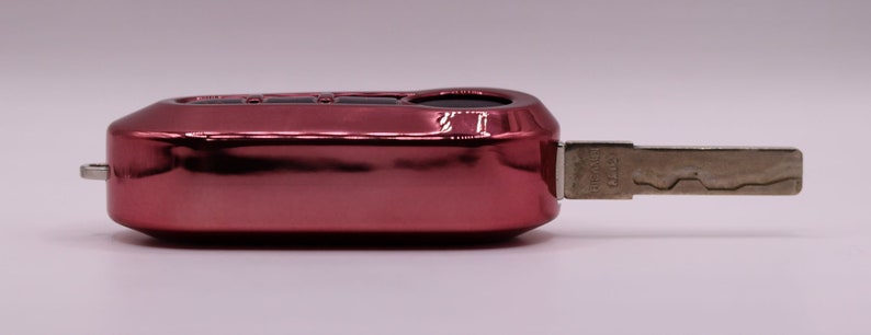 Puede incluir: Una llave de coche roja con una hoja de llave de metal plateado. La llave tiene un bot&oacute;n negro en la parte superior.