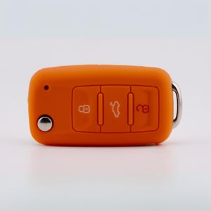 May include: A bright orange silicone car key fob cover featuring three embossed button icons for locking, unlocking, and opening the trunk. The cover has a small circular cutout for a key blade and a silver metal ring attached to the side.