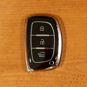 May include: A black and chrome car key fob featuring three buttons. The top button displays a locked padlock icon, the middle button shows an unlocked padlock icon, and the bottom button depicts a car outline with the text HOLD underneath it.