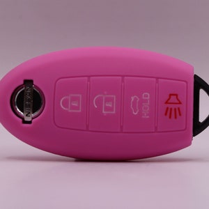May include: A bright pink car key fob cover with a black loop. The key fob has four buttons with white icons for lock, unlock, panic, and trunk release. The Nissan logo is visible on the key.