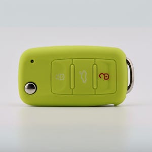 May include: A lime green silicone car key fob cover featuring three rectangular buttons with white and red icons for locking, unlocking, and trunk release, along with a circular silver metal button and a silver metal keyring attachment.