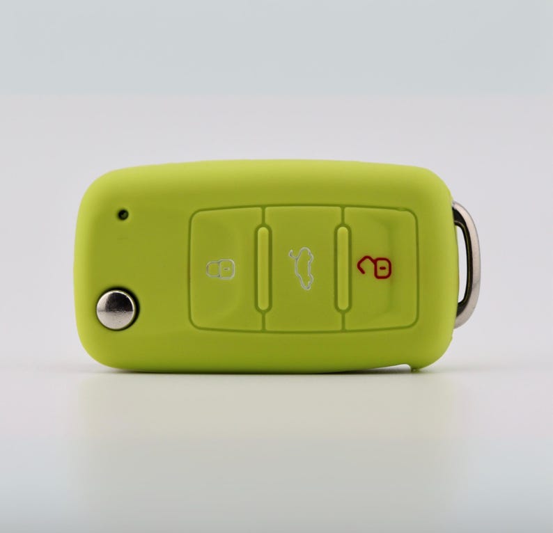 May include: A lime green silicone car key fob cover featuring three rectangular buttons with white and red icons for locking, unlocking, and trunk release, along with a circular silver metal button and a silver metal keyring attachment.