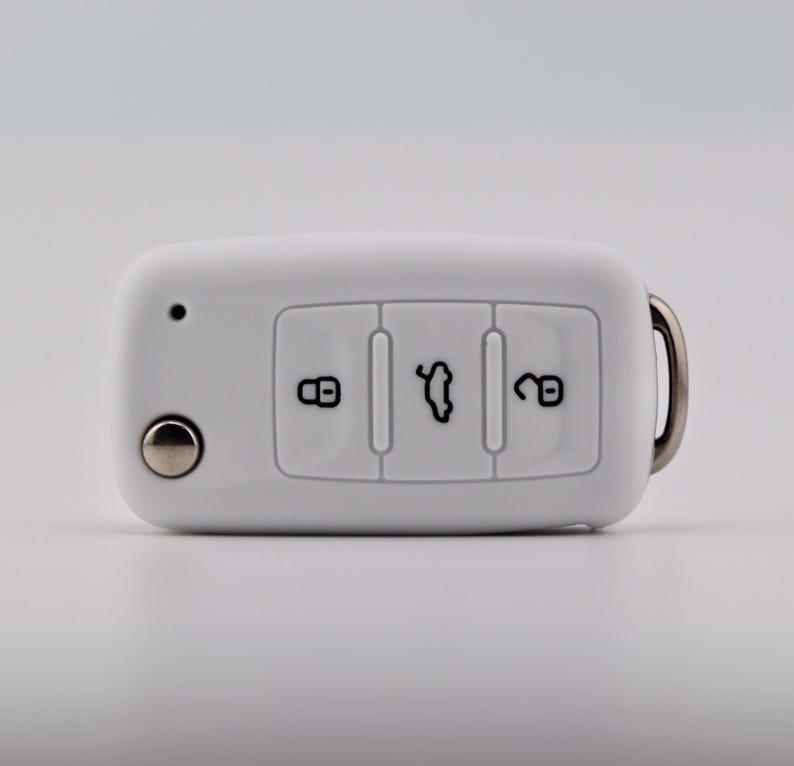 May include: A white rectangular car key fob featuring three buttons with black icons representing a locked padlock, an open trunk, and an unlocked padlock. A small circular silver button is on the left, and a metal keyring loop is on the right.