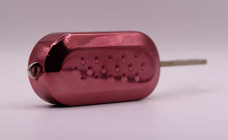 Puede incluir: Una llave de coche roja con una hoja de llave plateada. La llave tiene una superficie texturizada con peque&ntilde;as hendiduras circulares.