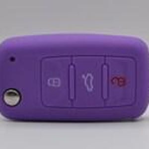 May include: A purple silicone car key fob cover featuring three rectangular buttons with white icons for locking, trunk release, and unlocking, along with a circular silver button and a metal ring attachment on the right side of the rectangular casing.