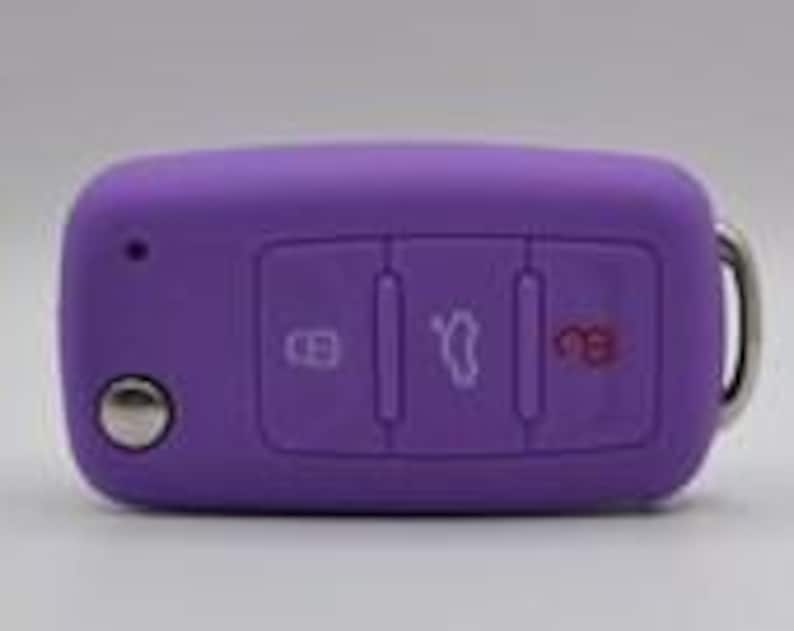 May include: A purple silicone car key fob cover featuring three rectangular buttons with white icons for locking, trunk release, and unlocking, along with a circular silver button and a metal ring attachment on the right side of the rectangular casing.